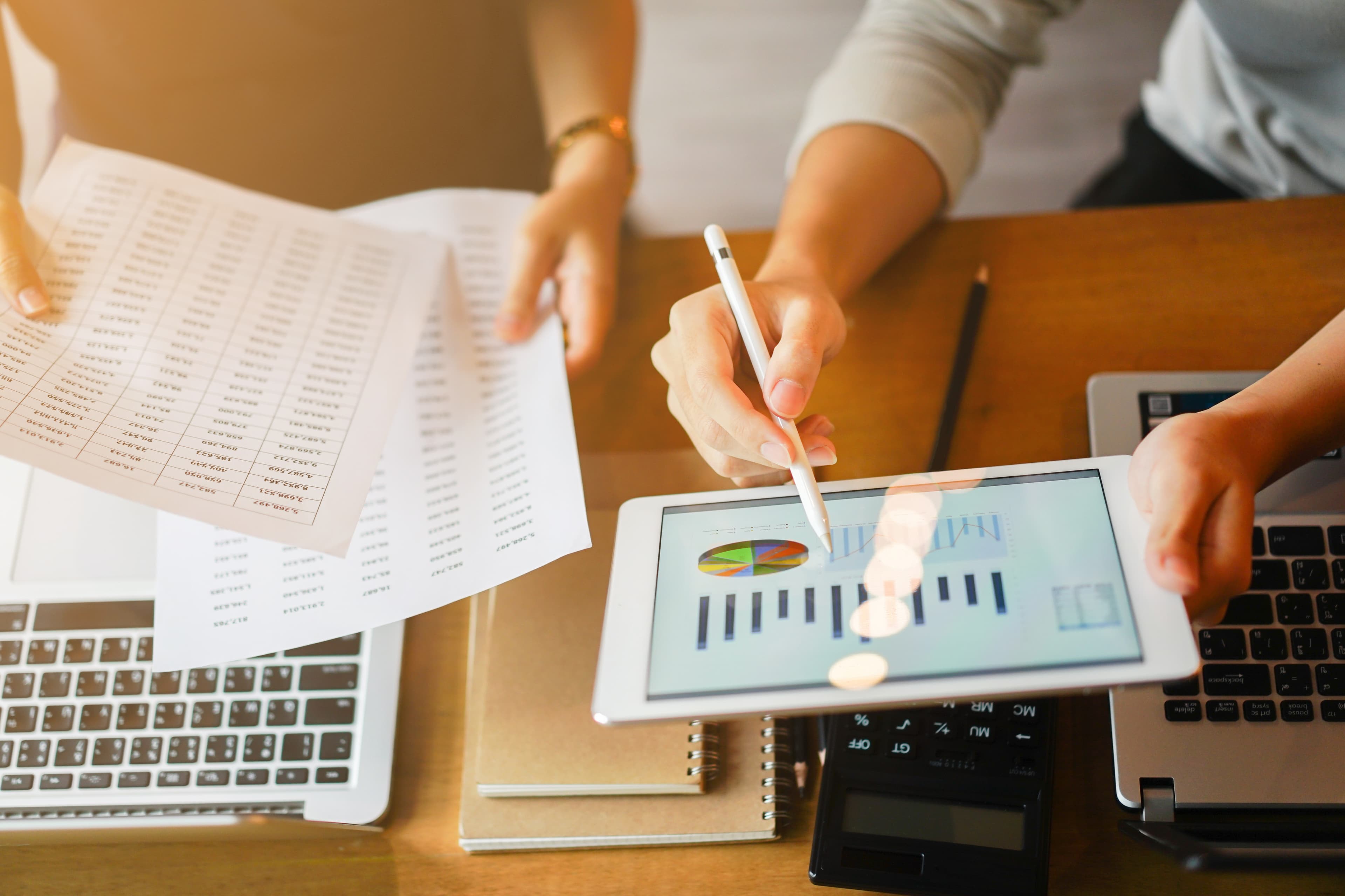 Two people reviewing financial data, with one person holding printed sheets and the other using a tablet with charts. Laptops and a calculator are on the table